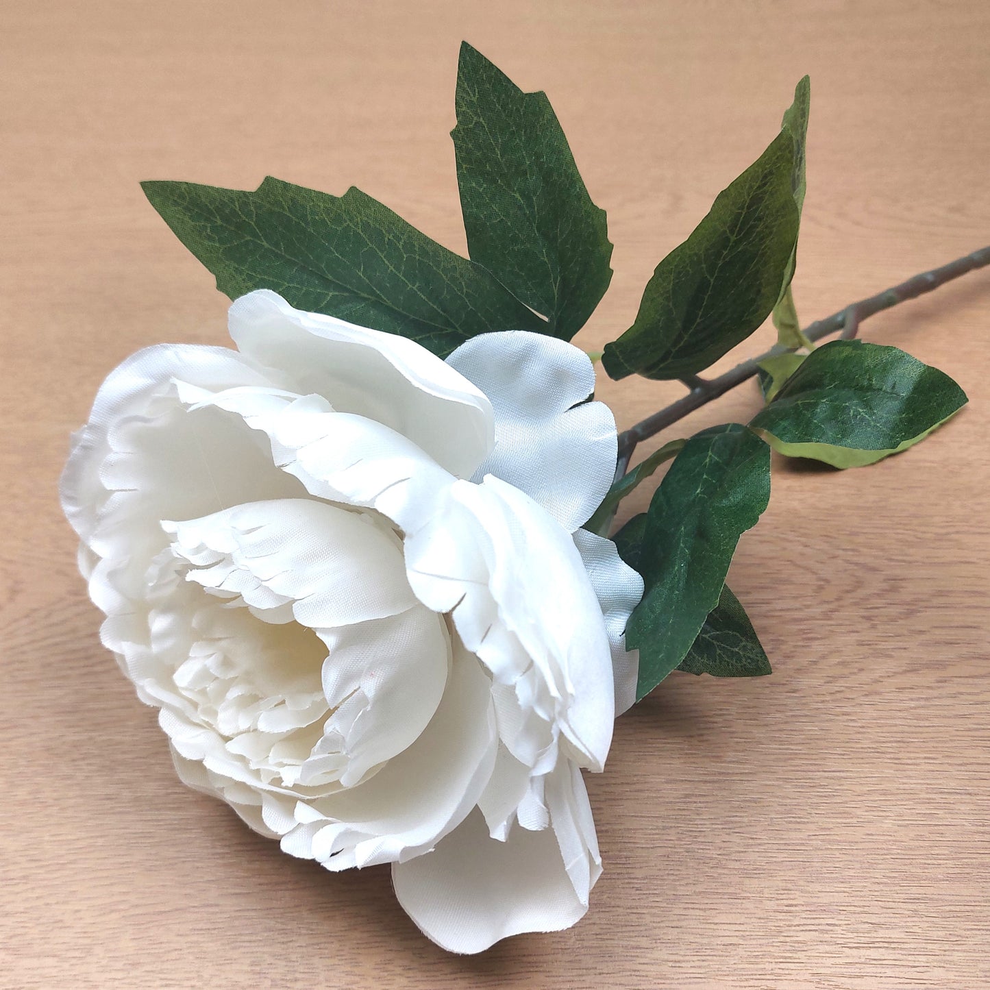 White artificial Peony flower with green leaves on a wooden surface