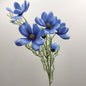 A spray of artificial blue Cosmos flowers with green stems and leaves, photographed against a plain light gray background.