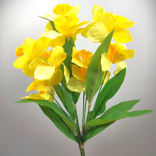A bunch of artificial Daffodils which are yellow flowers and green leaves against a pale background