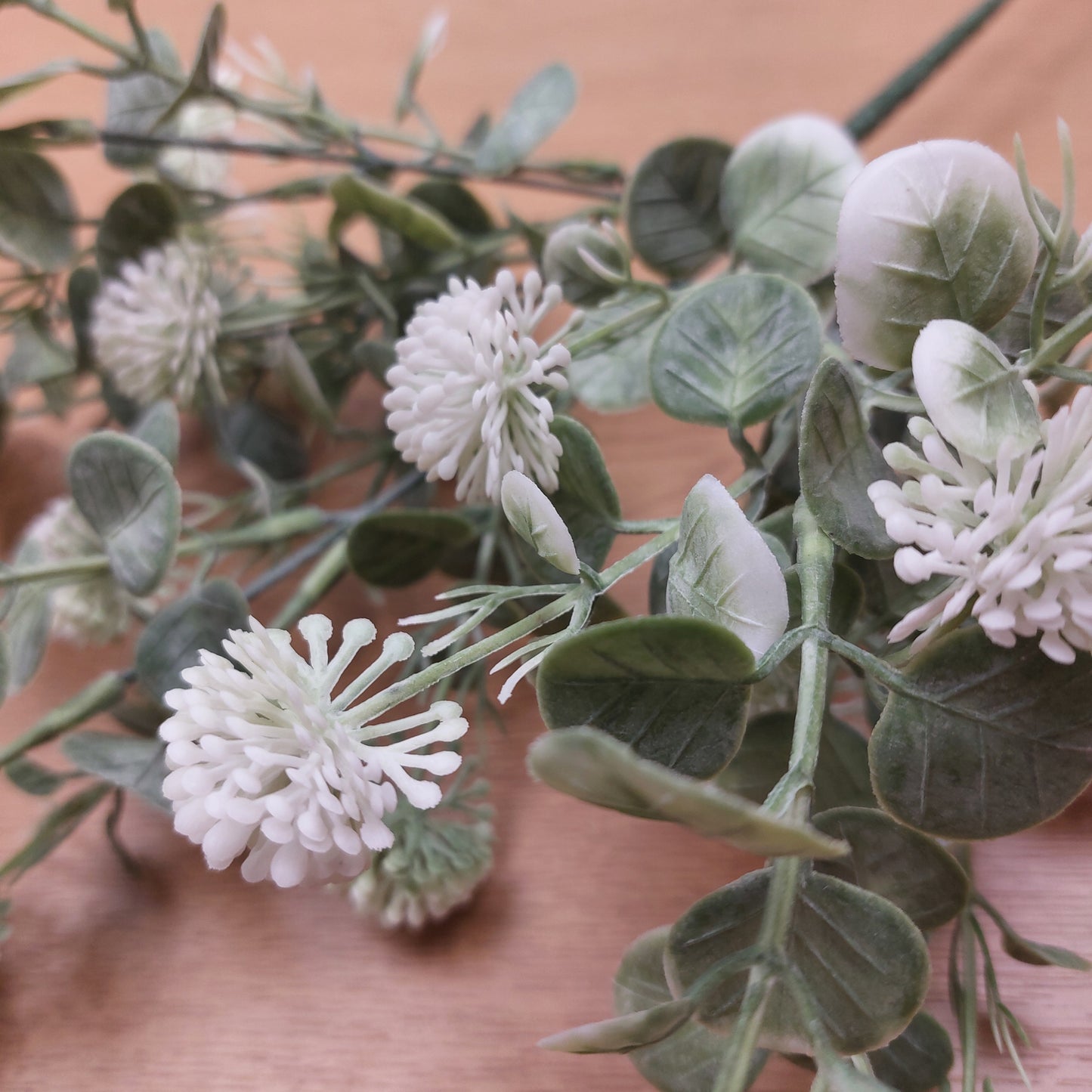 Artificial green foliage and white and green pom-pom flowers on a light wood background.