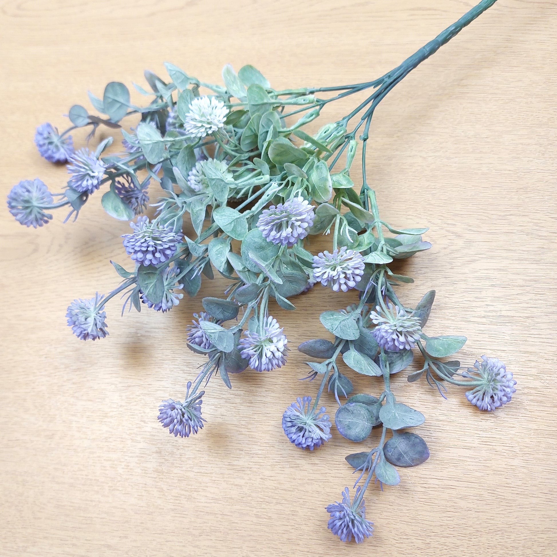 Artificial floral stem with dusty blue-purple globe flowers and muted green-gray leaves, arranged on a light wood surface.