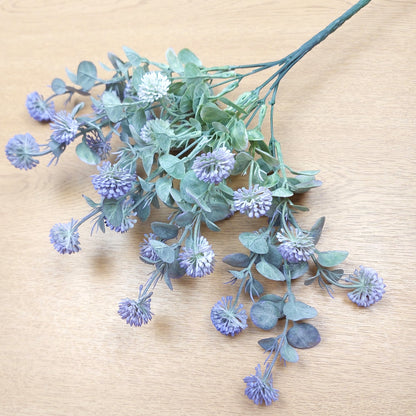 Artificial floral stem with dusty blue-purple globe flowers and muted green-gray leaves, arranged on a light wood surface.