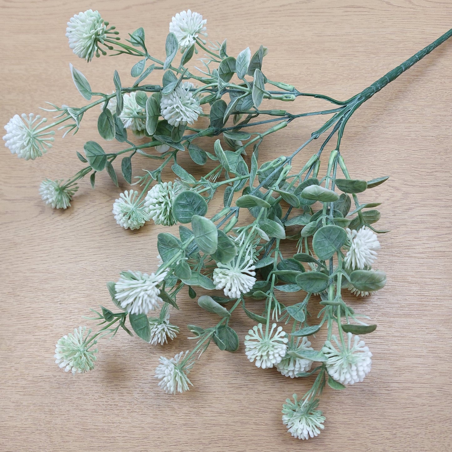 Artificial greenery stem with dusty green oval leaves and small white and green globular flower Echinops style clusters, laid on a light wood surface.