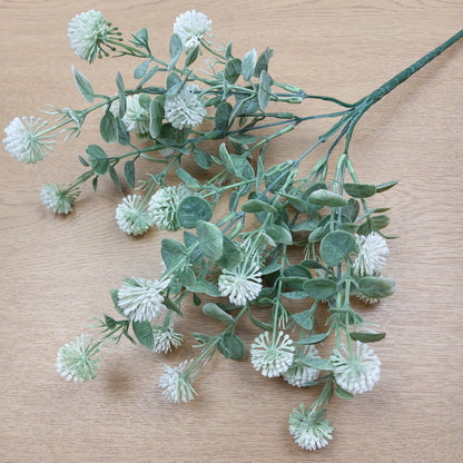 Artificial greenery stem with dusty green oval leaves and small white and green globular flower Echinops style clusters, laid on a light wood surface.