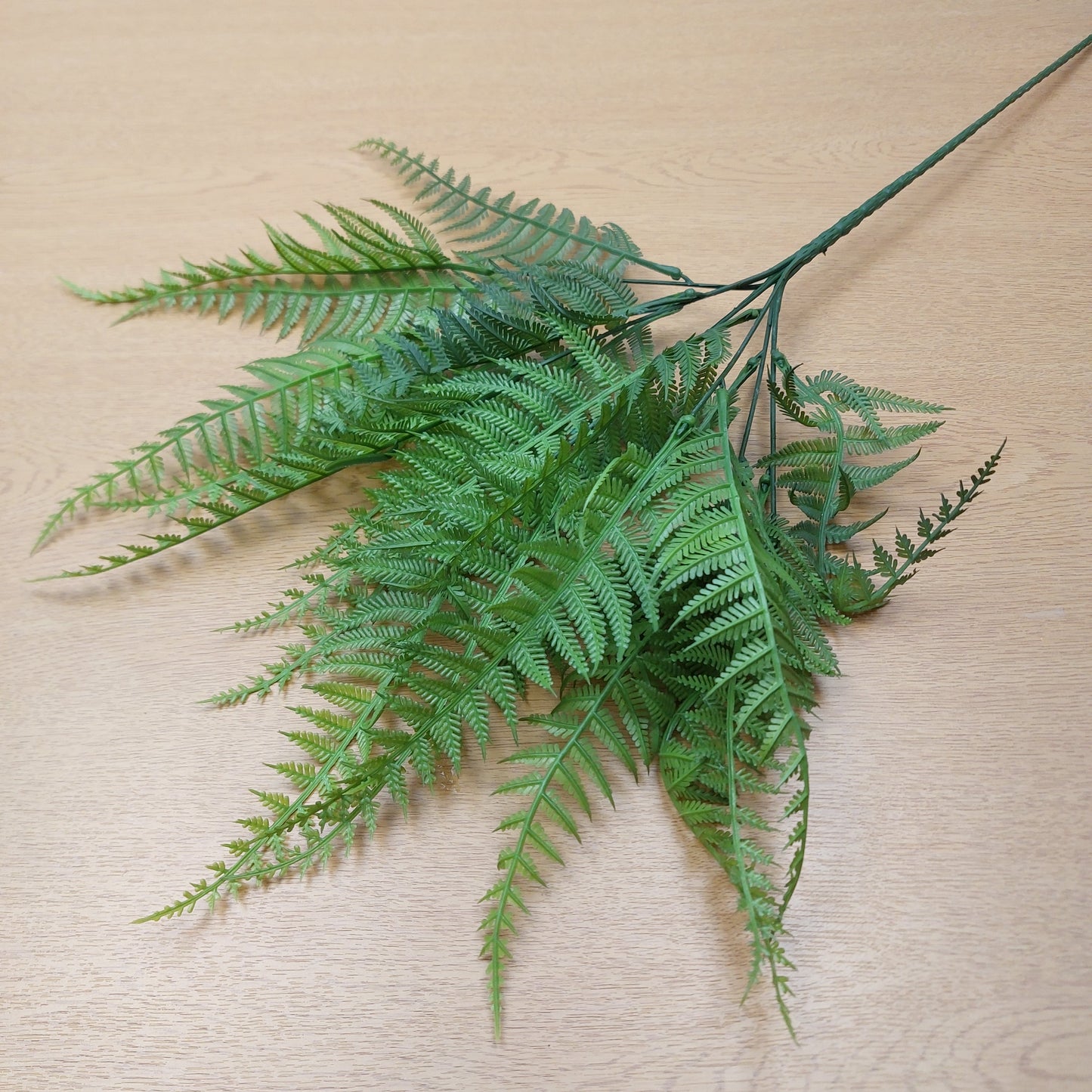 A single artificial fern branch with detailed green fronds, displayed against a light beige wood-grain background.