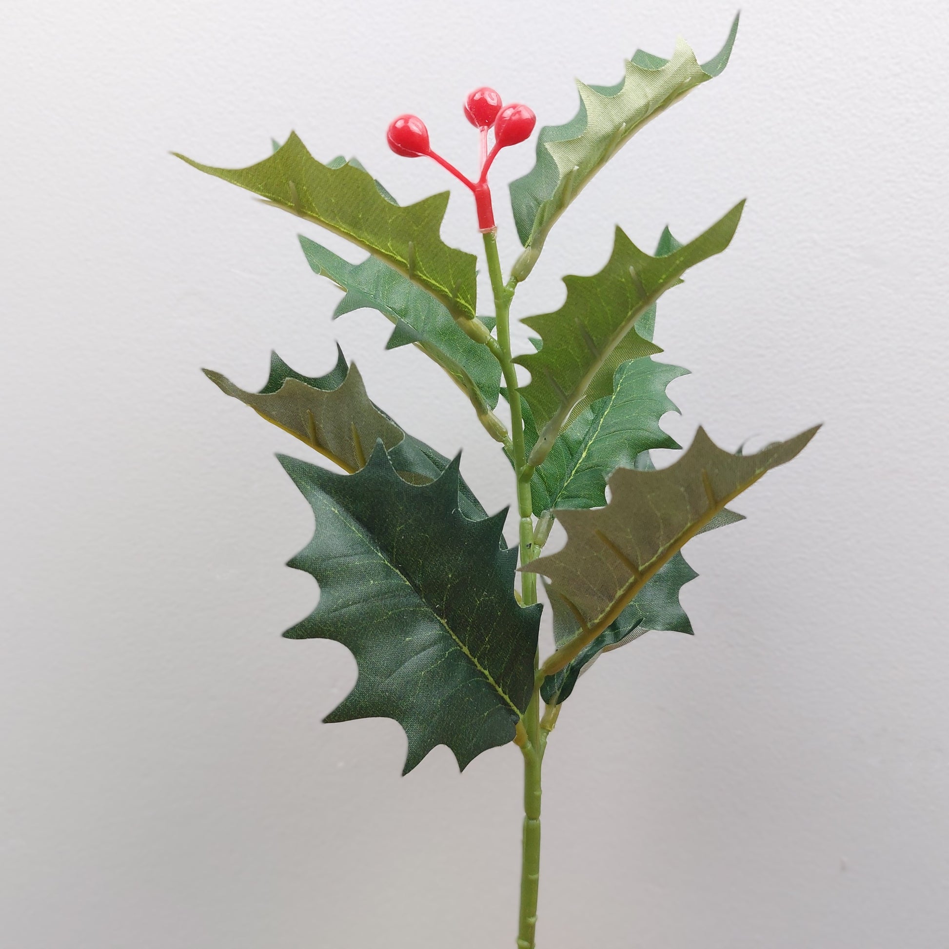Artificial Holly branch with green leaves and red berries on a light gray background