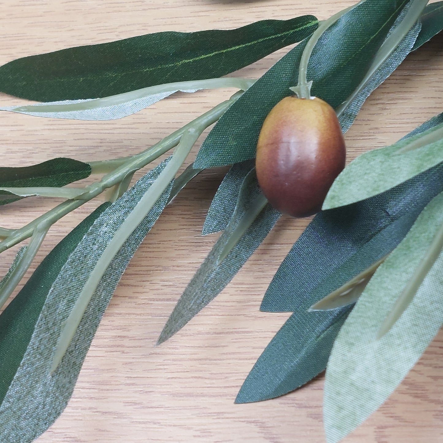 Close up of an Artificial olive branch with green leaves and a single brown olive on a wooden surface