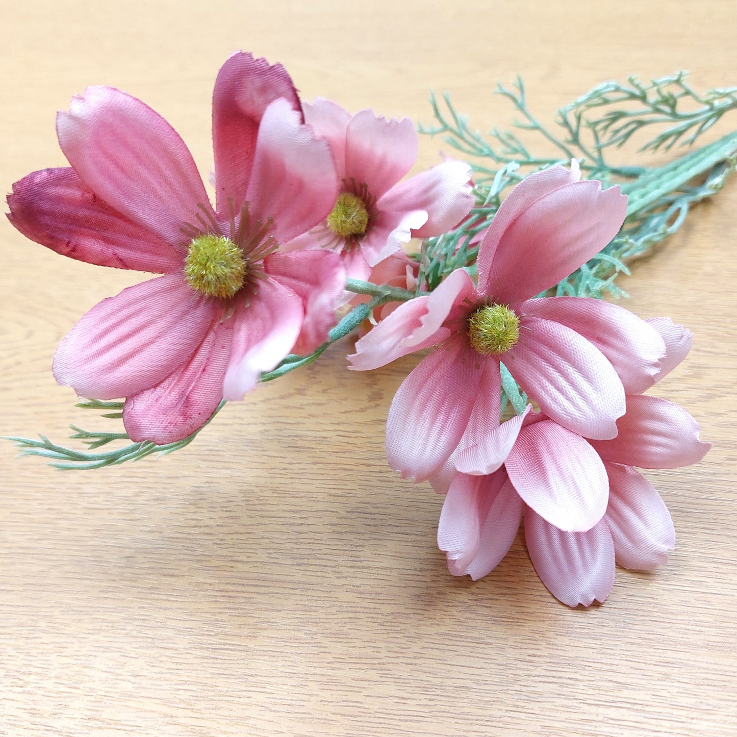 Pink artificial cosmos flowers with green stems and leaves, arranged on a light wood surface.