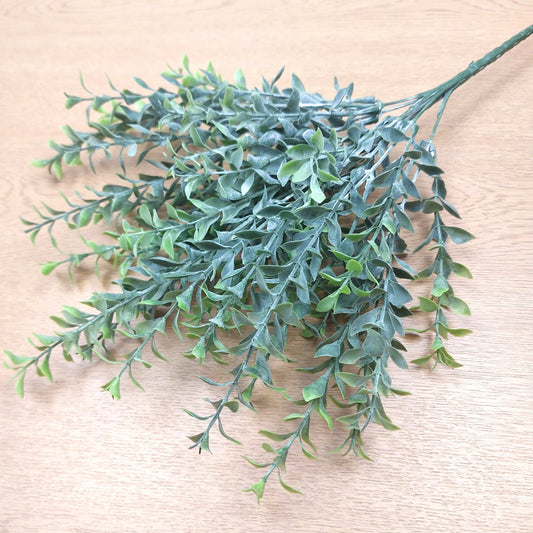 Artificial green leafy Parvifolia branch with dense small oval leaves on a light wood surface.