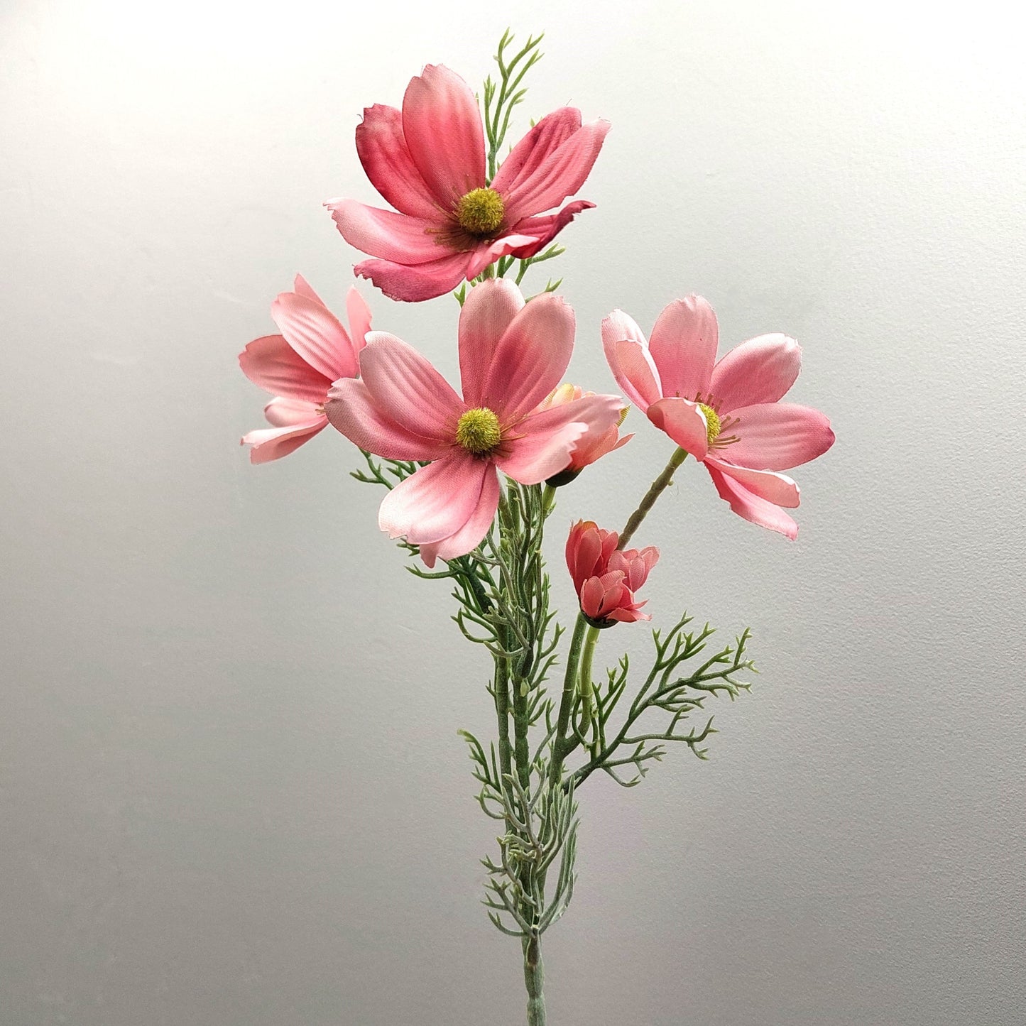 An artificial pink cosmos flower spray with green stems and feathery foliage, photographed against a plain light gray background.
