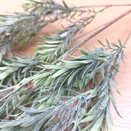 Close-up of artificial green raven leaf branches with needle-like leaves on a light wood surface.