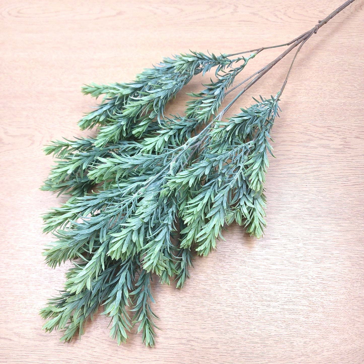Artificial green raven leaf spray with needle-like leaves on a light wood grain background.