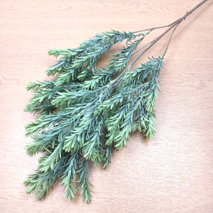 Artificial green raven leaf spray with needle-like leaves on a light wood grain background.