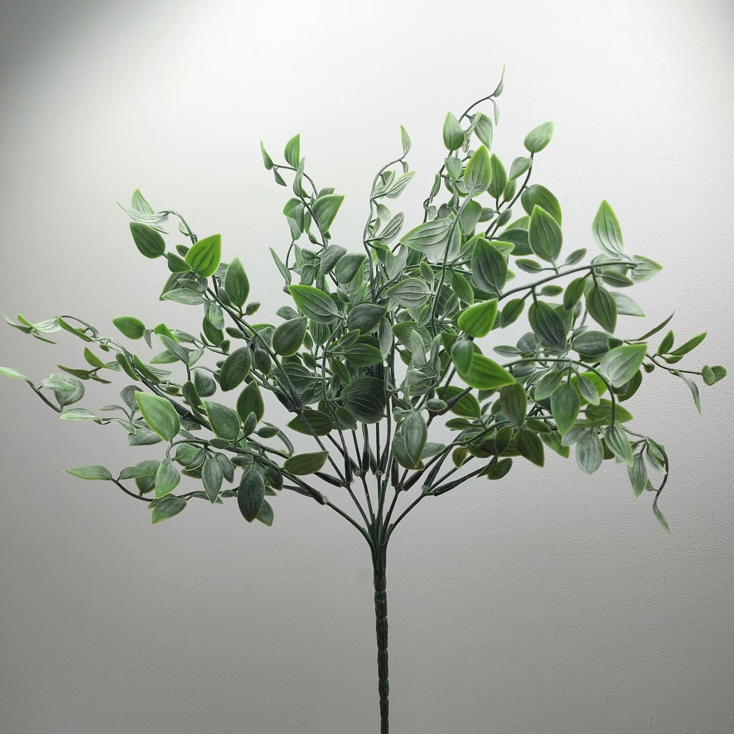 An artificial green leafy ruscus plant with multiple stems and oval-shaped leaves, shown against a plain light gray background.