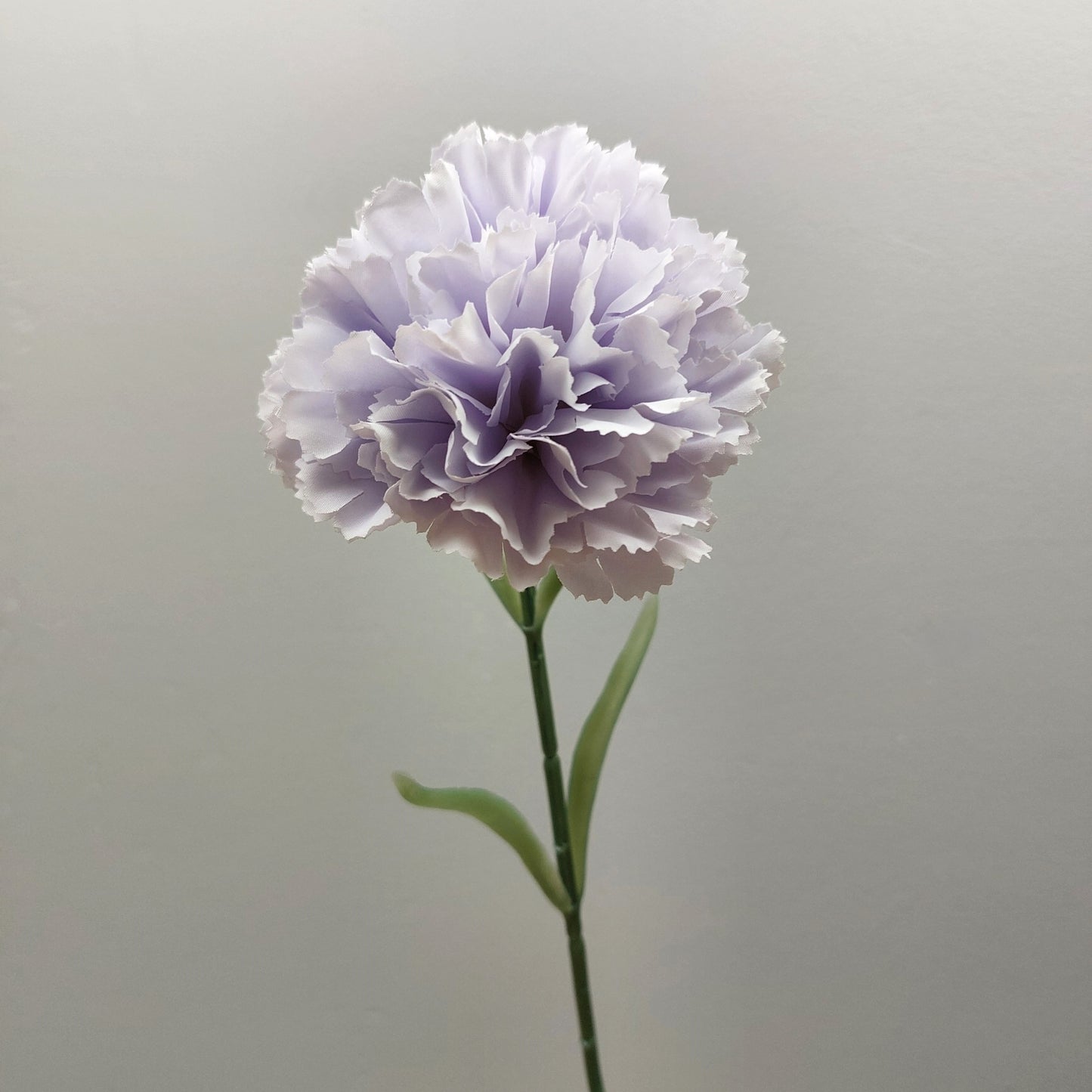 A single artificial lavender and lilac carnation flower with layered ruffled petals on a green stem, photographed against a plain light gray background.