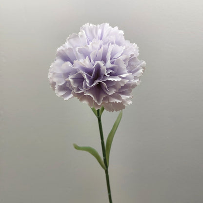 A single artificial lavender and lilac carnation flower with layered ruffled petals on a green stem, photographed against a plain light gray background.