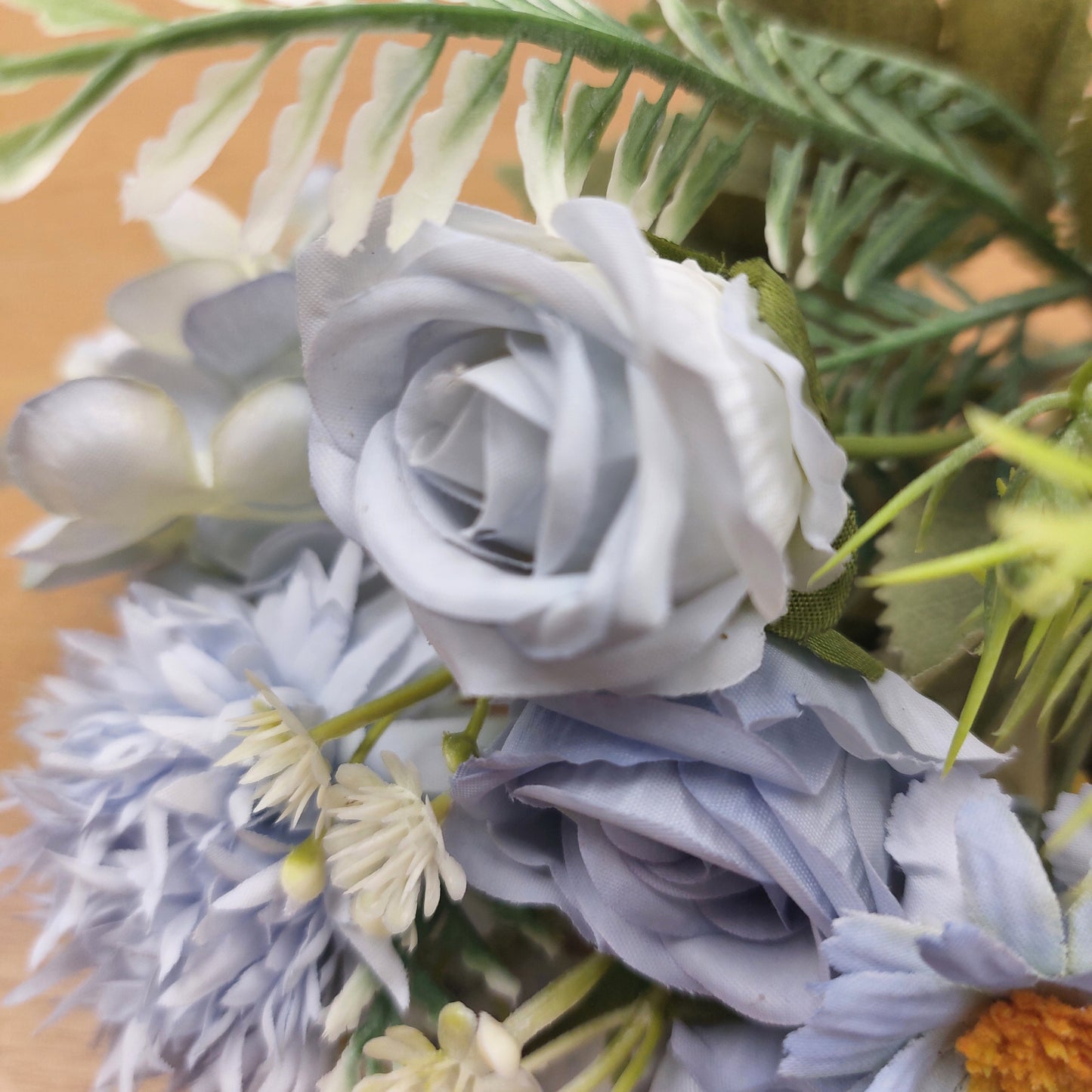 A close-up of a faux floral arrangement featuring pale blue and white roses, daisy-like flowers, and green leaves on a warm tan background.