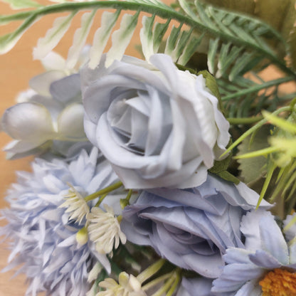 A close-up of a faux floral arrangement featuring pale blue and white roses, daisy-like flowers, and green leaves on a warm tan background.
