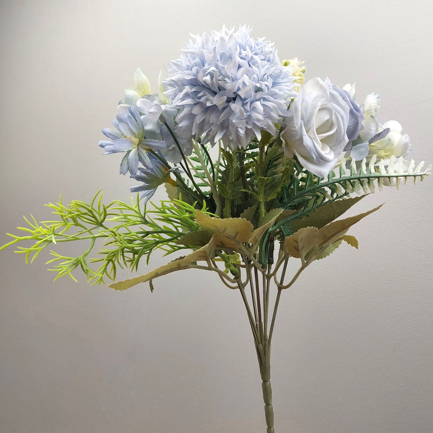 An artificial flower bouquet featuring light blue and white flowers with green and yellow foliage, set against a plain light gray background.