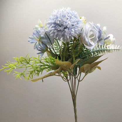 An artificial flower bouquet featuring light blue and white flowers with green and yellow foliage, set against a plain light gray background.