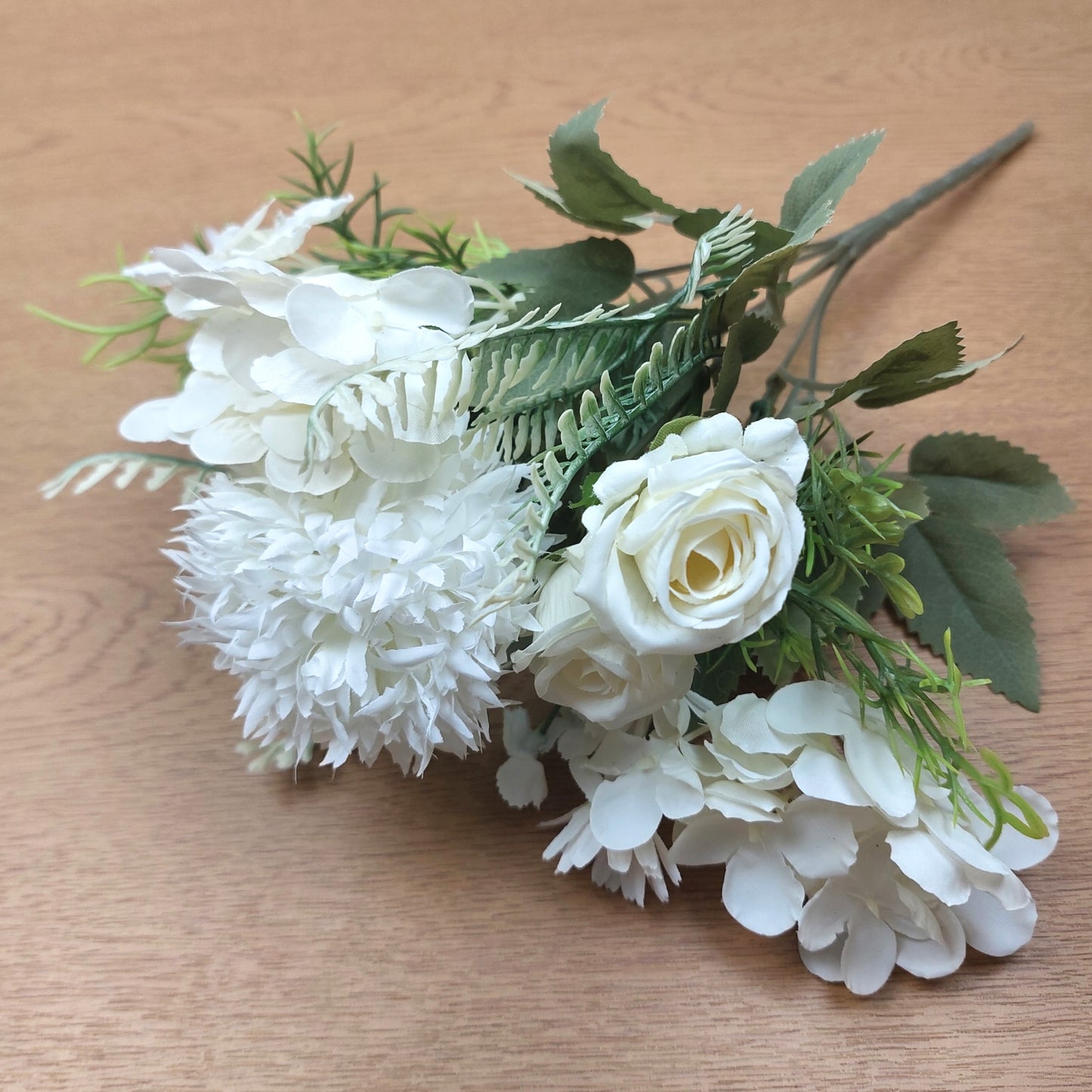 White artificial flower bouquet with white roses, white hydrangeas, and green leaves on a light brown wooden surface.