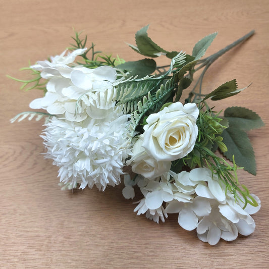 White artificial flower bouquet with white roses, white hydrangeas, and green leaves on a light brown wooden surface.