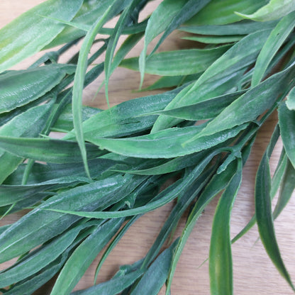 Green artificial wild grass-like leaves with a realistic textured finish, arranged on a light wood surface.