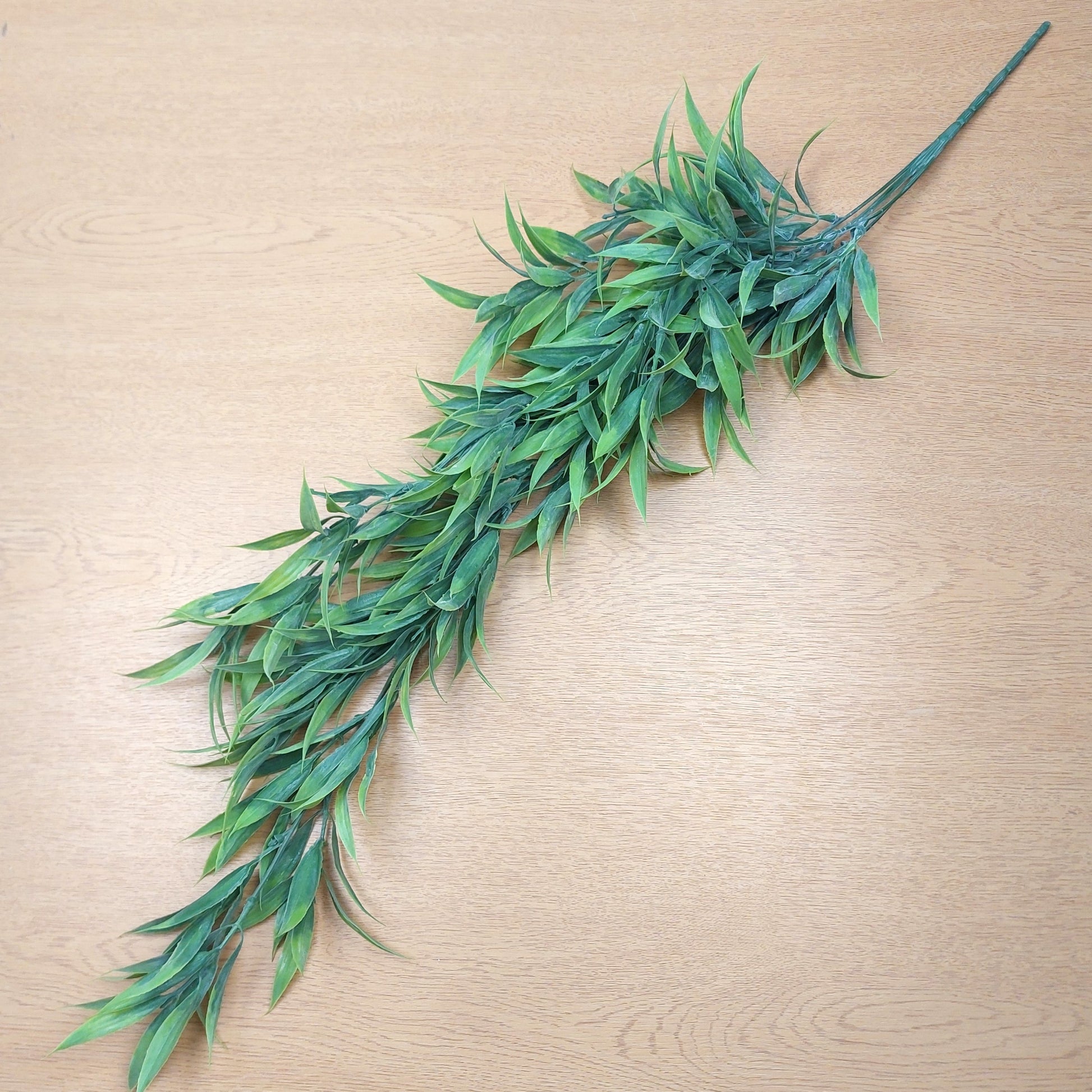 A long artificial green leafy trailing wild grass with many narrow leaves and a long stem, laid diagonally on a light wood surface.