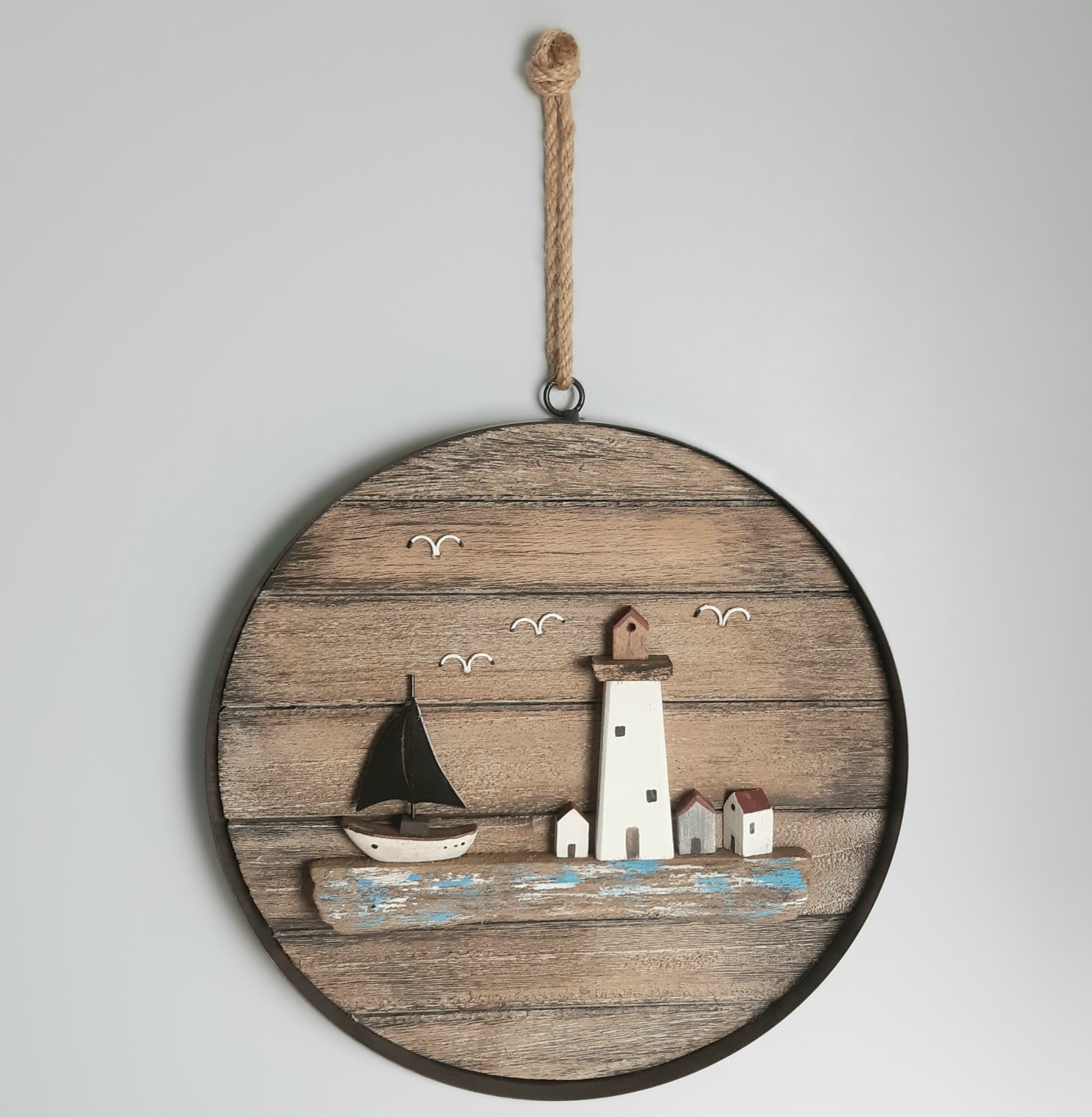 Decorative wall art with a lighthouse and sailboat on a wooden circle, hanging against a light gray background.