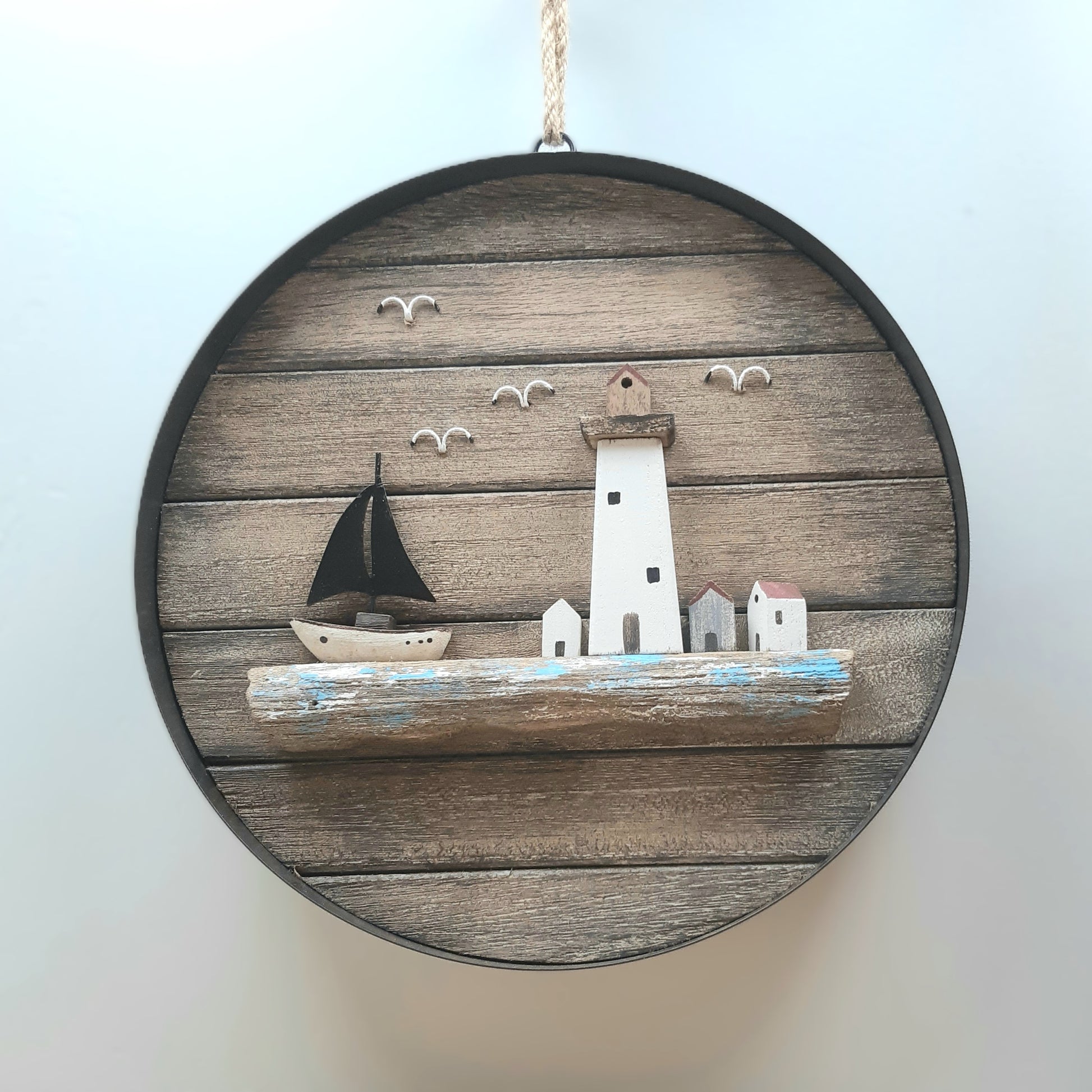 A wooden wall hanging depicting a scene of a boat with lighthouse and buildings with 4 birds and the sea, surrounded by a metal ring and hanging by a knotted rope.