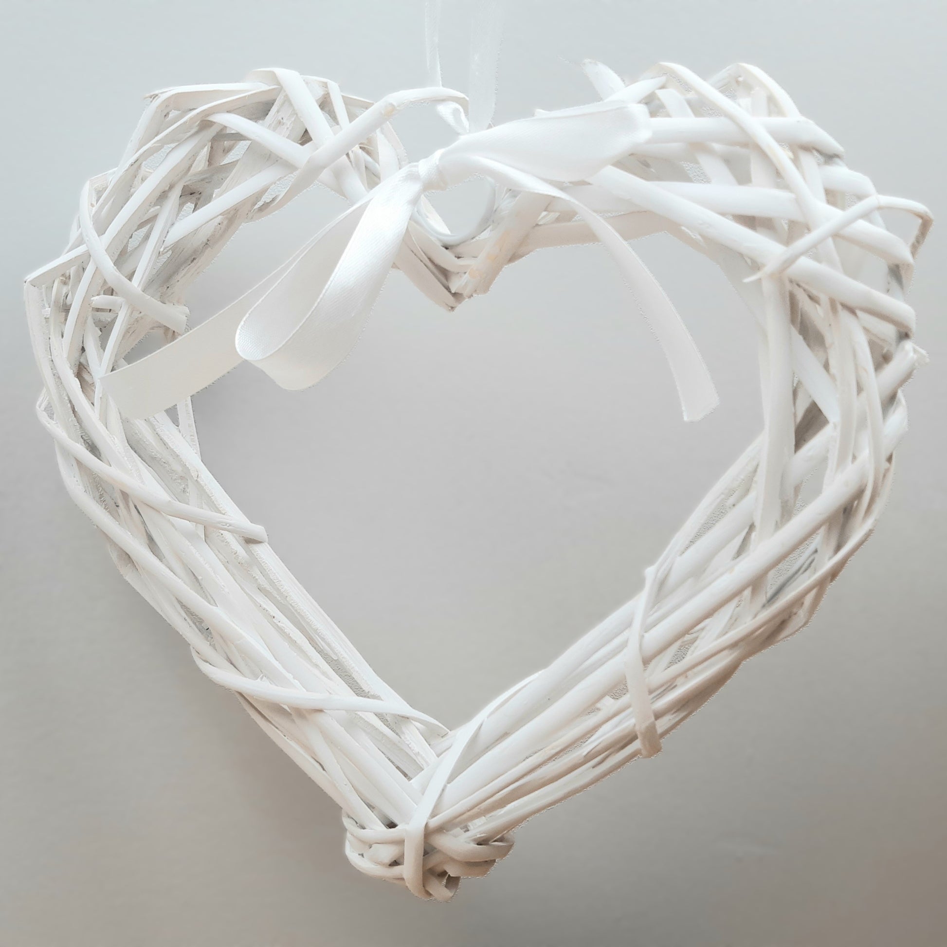 A hanging White Colour Wicker Heart with a white satin ribbon and bow.