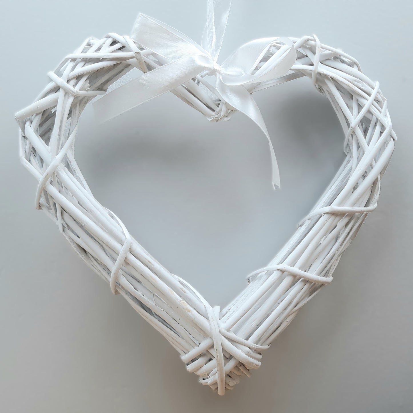 A hanging White Colour Wicker Heart with a white satin ribbon and bow.