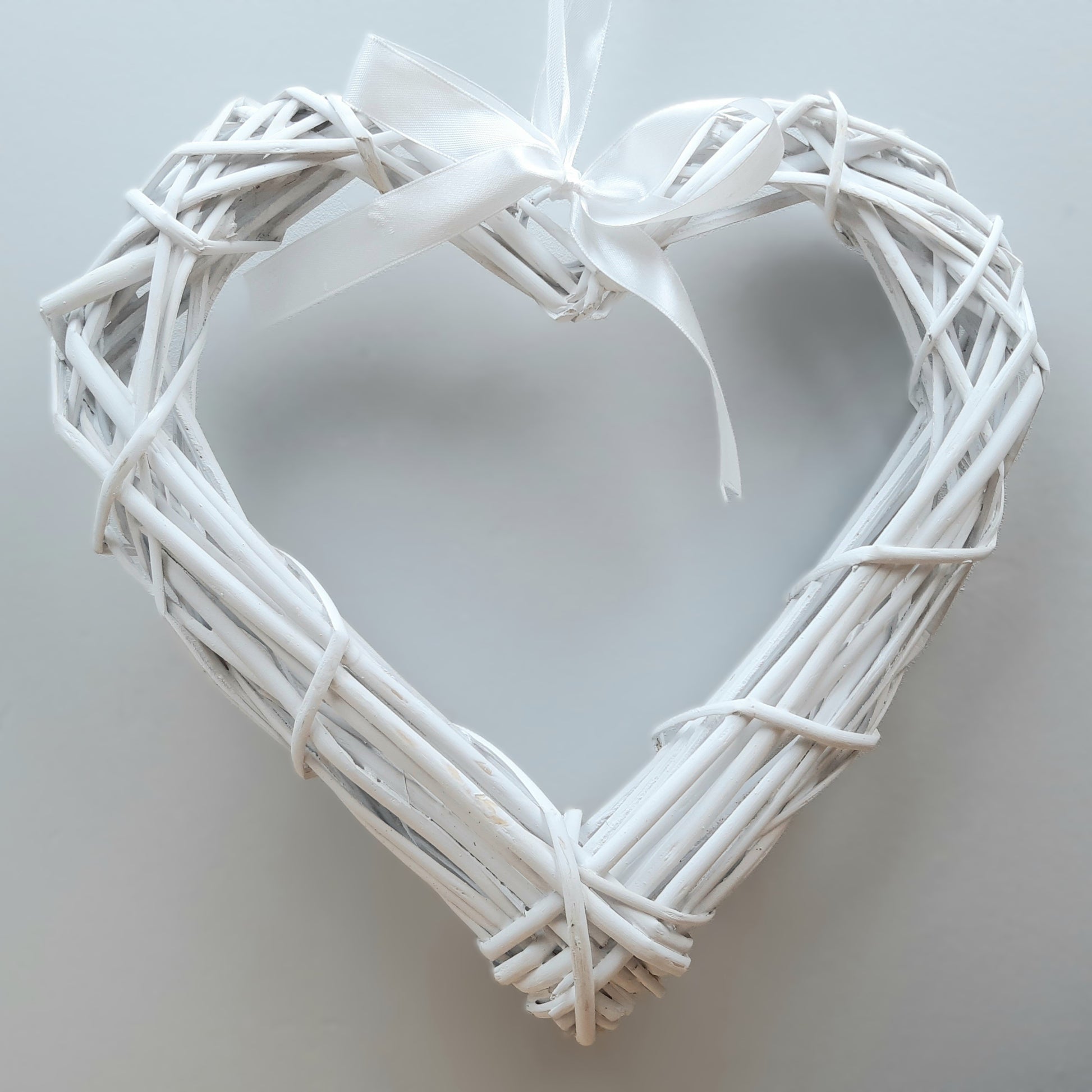 A hanging White Colour Wicker Heart with a white satin ribbon and bow.