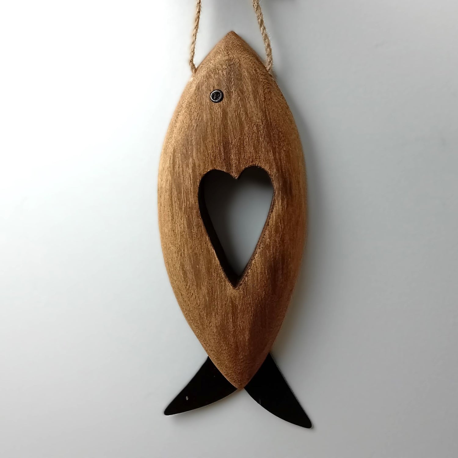 A brown wooden carved fish with metal fins and eye, with a heart shape carved in the middle hanging from a jute rope against a pale background.