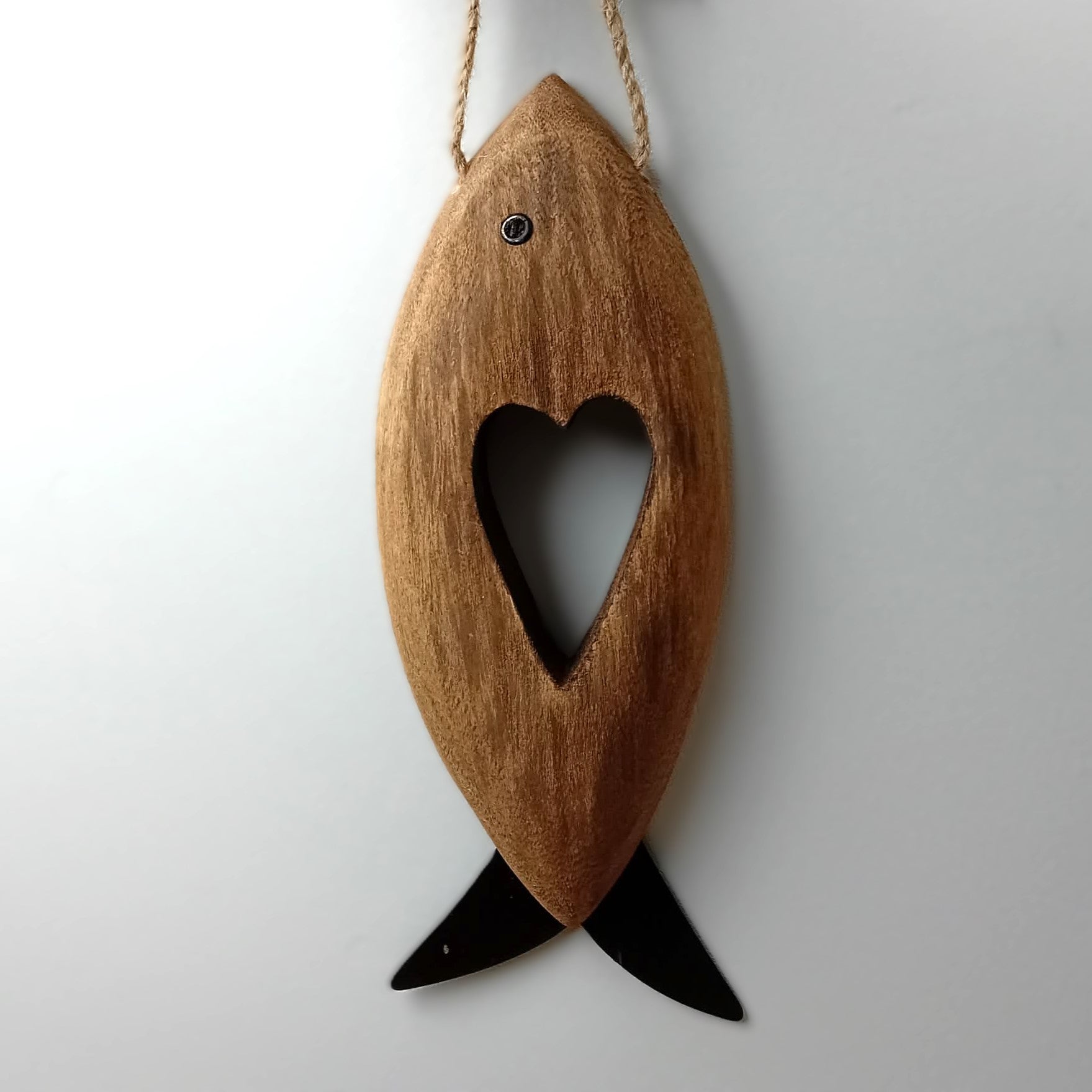 A brown wooden carved fish with metal fins and eye, with a heart shape carved in the middle hanging from a jute rope against a pale background.