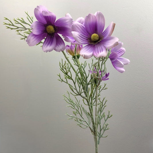 Artificial Purple cosmos-style flowers with Lilac to lavender petals and yellow centers, arranged with green feathery stems against a plain light gray background.