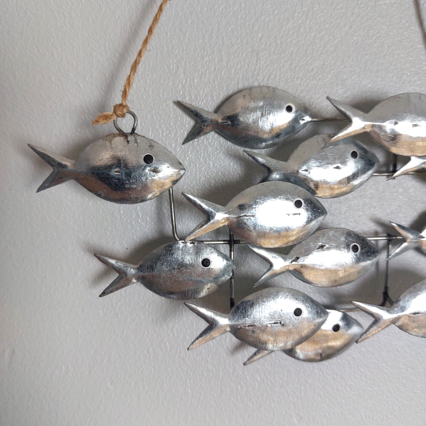 Tin metal fish wall hanging decor featuring multiple small silver fish suspended by thin metal wires and a natural jute hanging string, shown against a plain light gray wall.