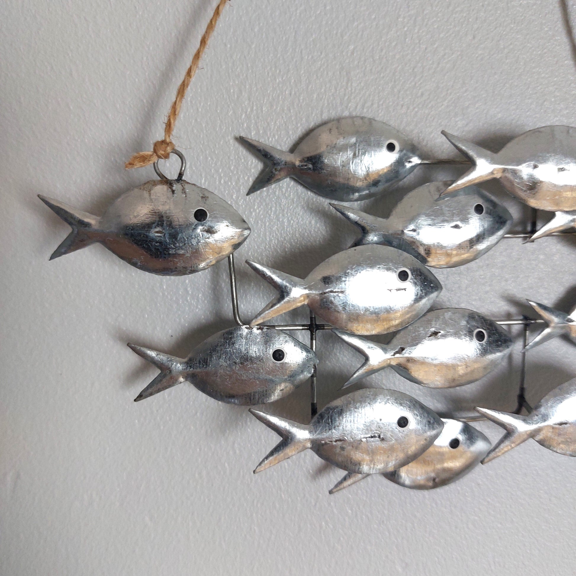 Tin metal fish wall hanging decor featuring multiple small silver fish suspended by thin metal wires and a natural jute hanging string, shown against a plain light gray wall.