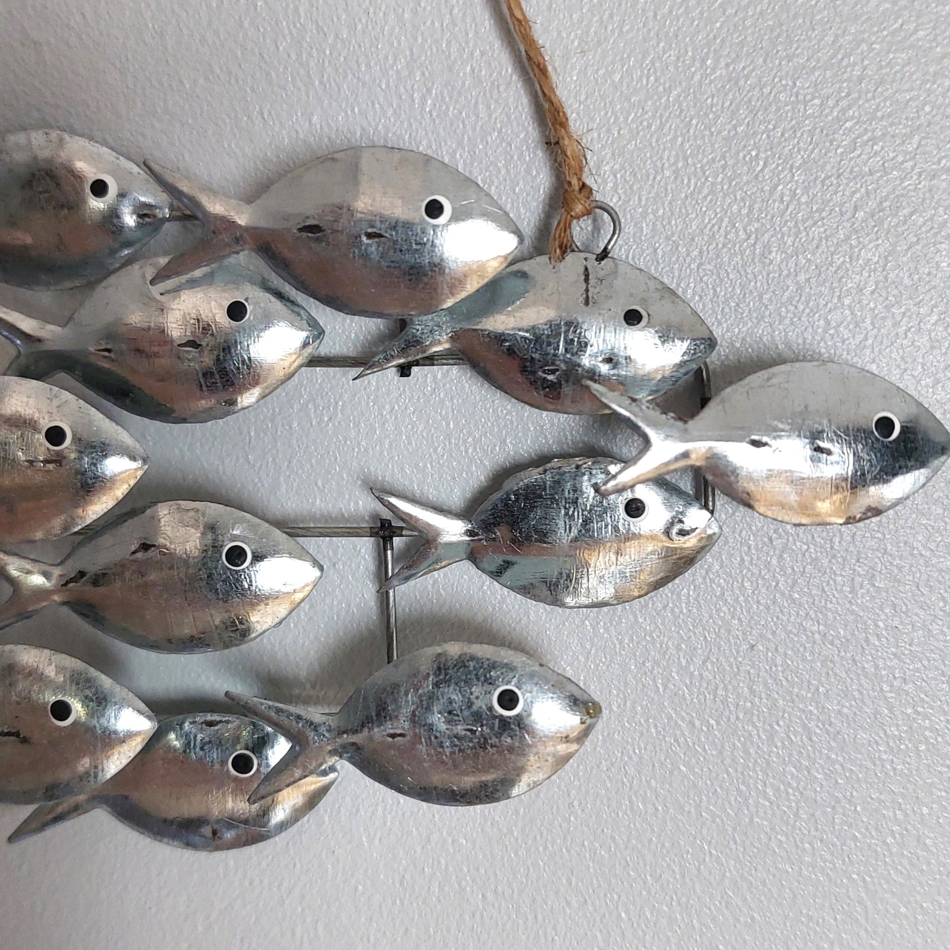 A decorative wall hanging made of multiple small silver fish shapes attached to a metal frame, with a natural jute hanging twine at the top, shown against a light gray textured wall.