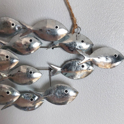 A decorative wall hanging made of multiple small silver fish shapes attached to a metal frame, with a natural jute hanging twine at the top, shown against a light gray textured wall.