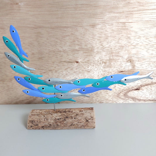 A decorative hand painted metal fish sculpture depicting a school of small blue, teal, and silver fish, mounted on a rustic wooden base against a light wood panel background.