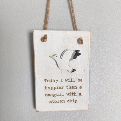 A whitewashed style wooden plaque, showing a seagull with the phrase "Today I will be happier than a seagull with a stolen chip" hanging from a jute string against a pale background.