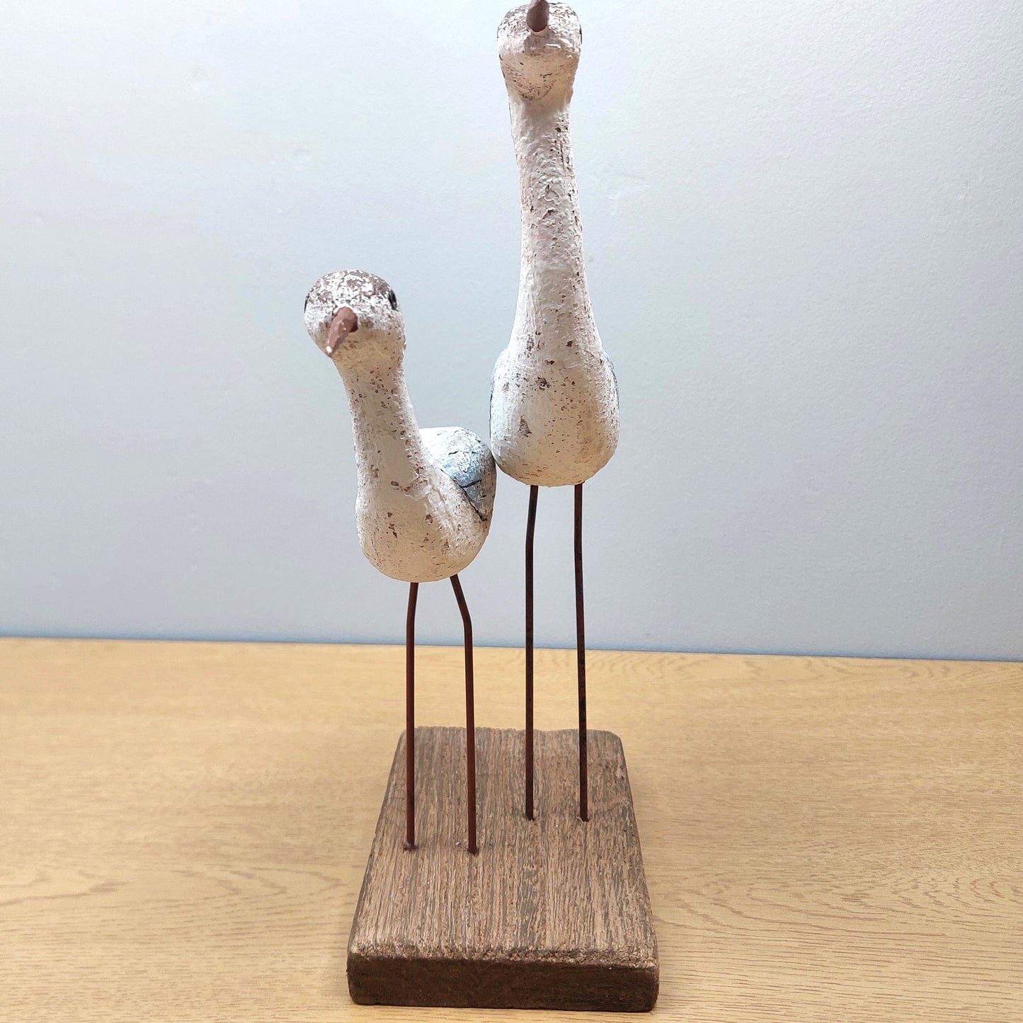 Two wooden wading birds facing head on, on a wooden base against a light blue background