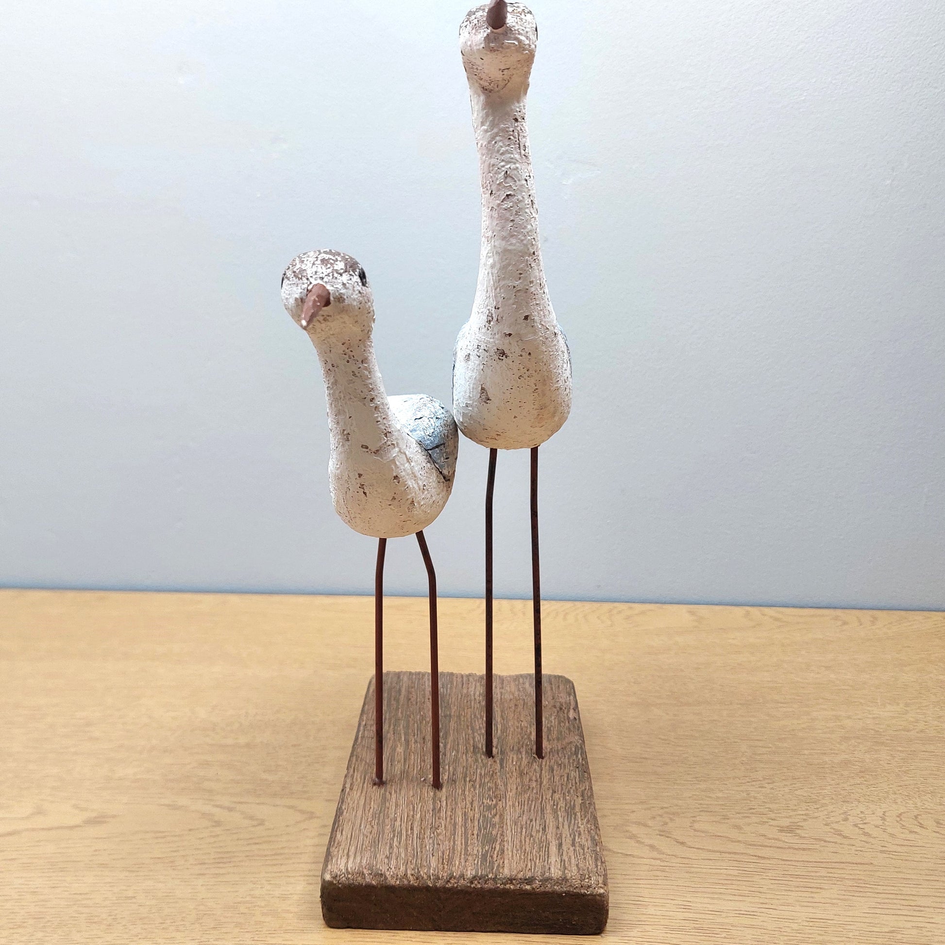 Two wooden wading birds facing head on, on a wooden base against a light blue background