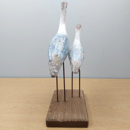 Two wooden wading birds facing away on a wooden stand against a gray background
