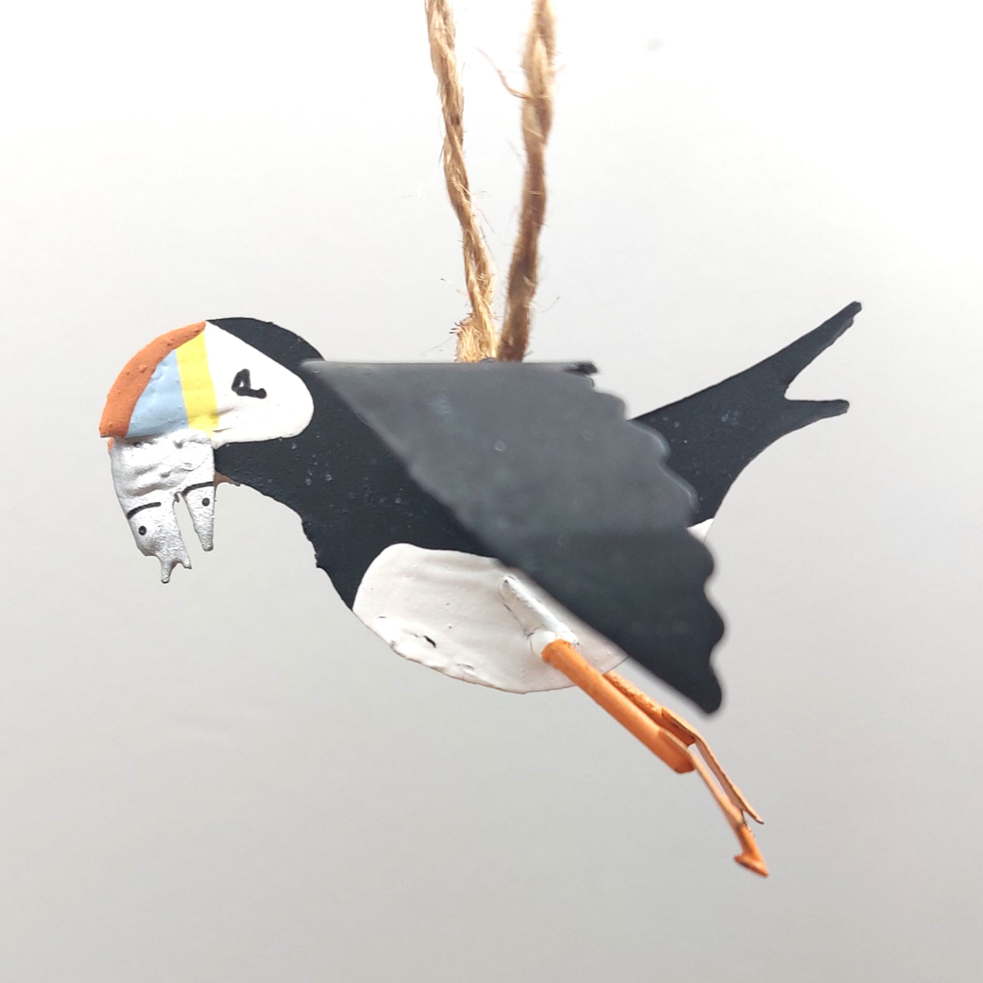 Decorative metal ornament shaped like a puffin carrying some sand eels hanging from a jute string on a white background