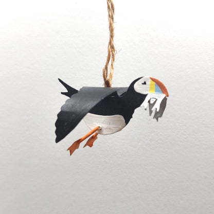 A metal hand painted puffin carrying some sand eels away, mounted on a jute string against a pale background.