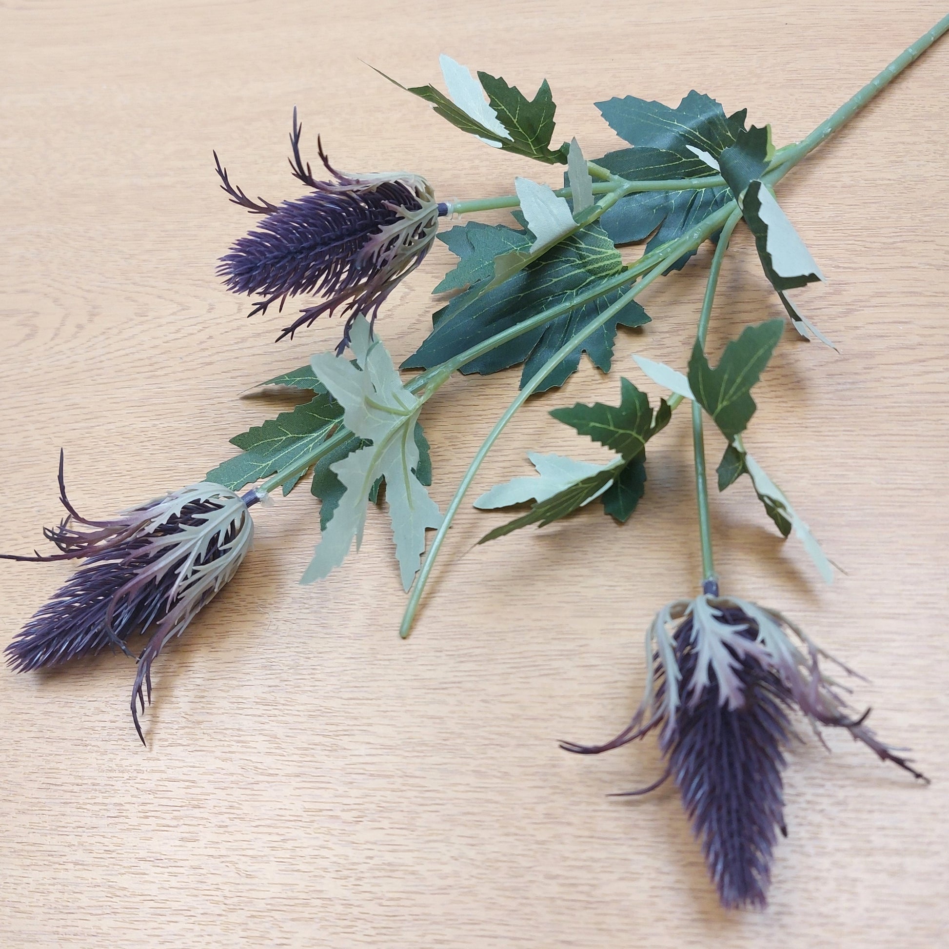 An artificial purple eryngium thistle spray with three heads and green leaves on stems, arranged on a light wood surface.