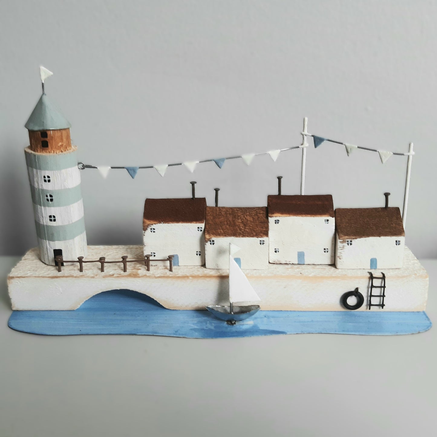 Model of a coastal scene with a lighthouse, houses, and a sailboat on a gray background
