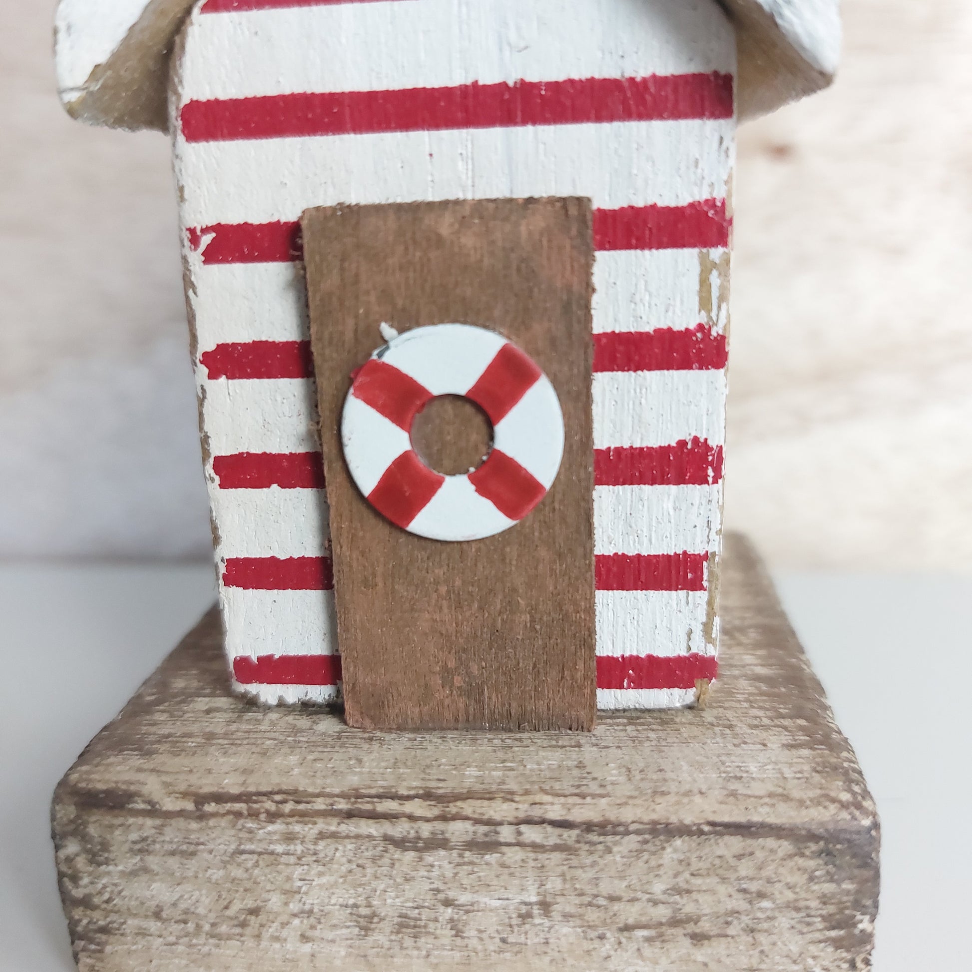 A small decorative wooden Beach Hut ornament with red and white horizontal stripes, and a red-and-white life preserver on the wooden brown door, displayed on a rustic wooden block against a soft neutral background.